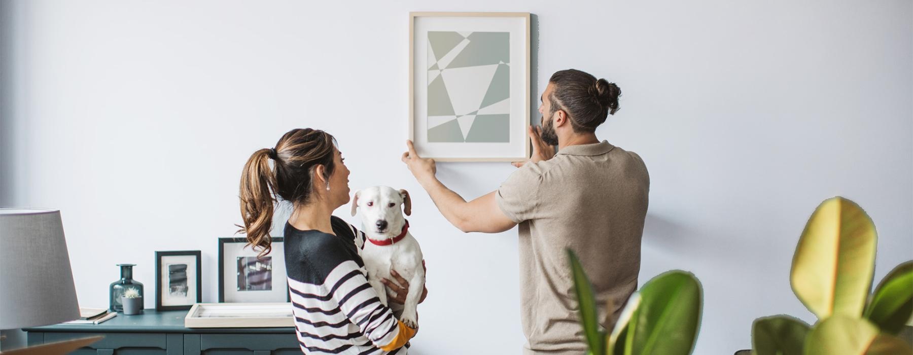 a man and a woman holding a dog while hanging a picture